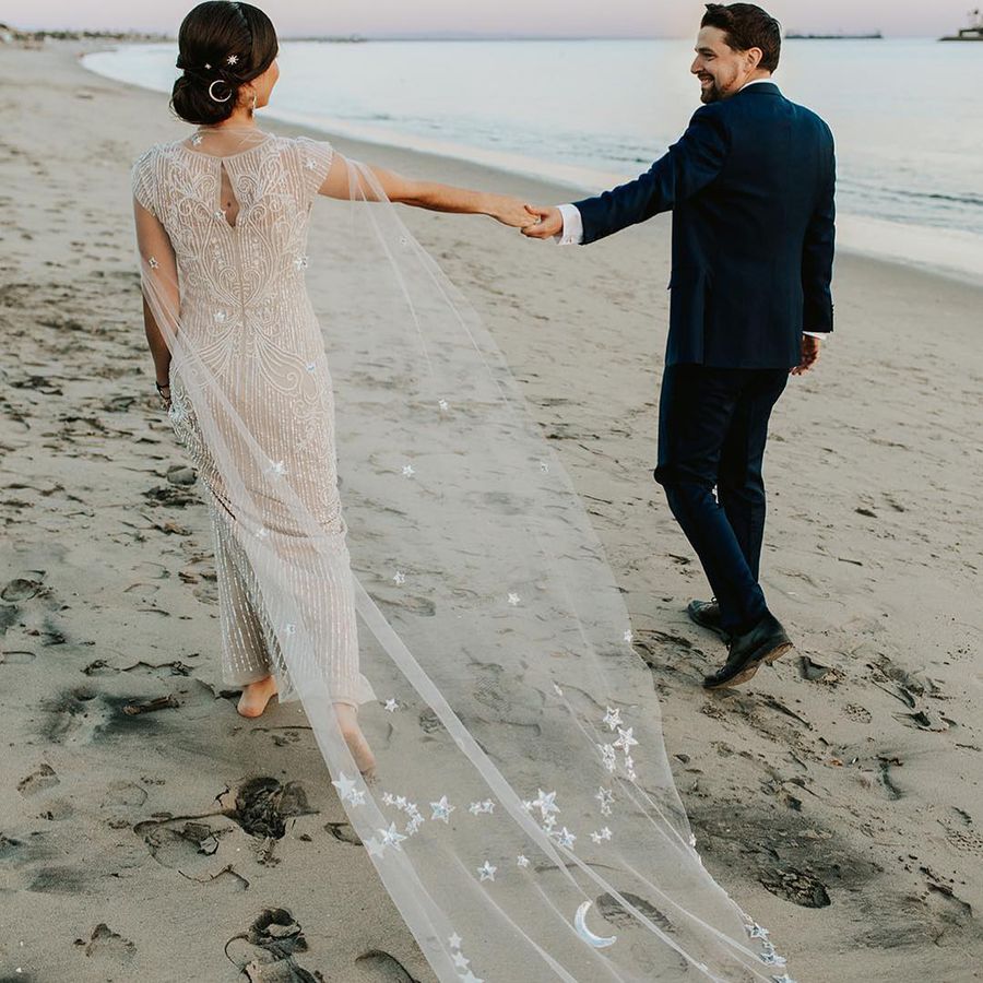 Bride in Celestial-Inspired Wedding Cape with Stars and Moons Holding Hands with Groom on the Beach