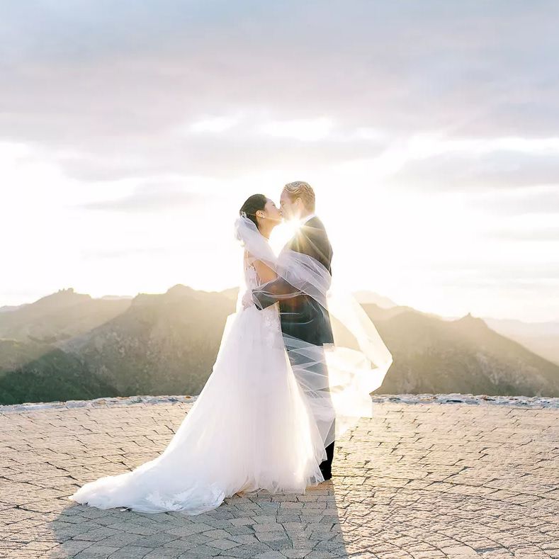 A bride and groom kissing with sunlight behind them and mountains in the distance.