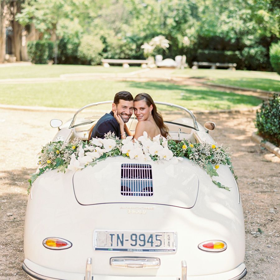 Bride and groom riding in the back of a vintage vehicle with orchids and greenery lining the hood