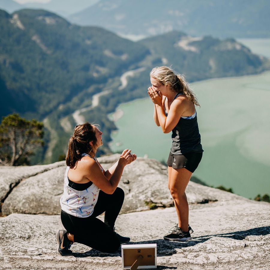 brunette woman proposing to blonde woman next to picture frame on top of a mountain overlooking a lake