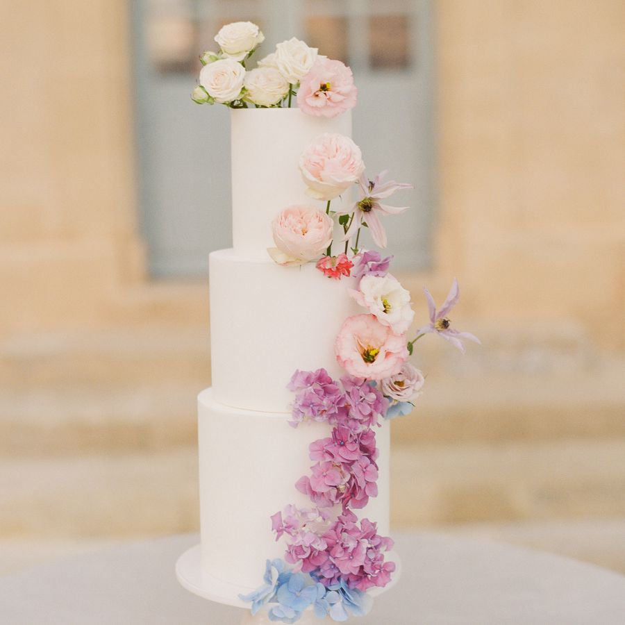 Three-tier white cake with purple and blue hydrangea and other colorful flowers