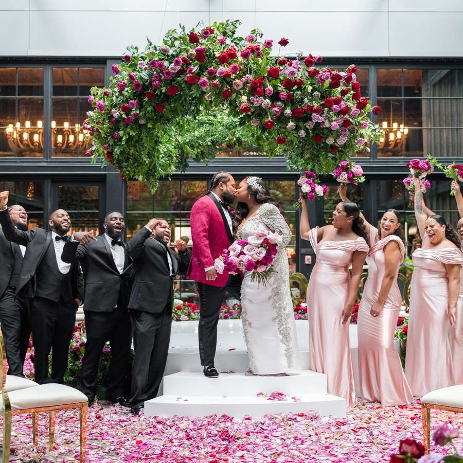 Couple kissing beneath a flower chandelier of pink and red roses with their wedding party