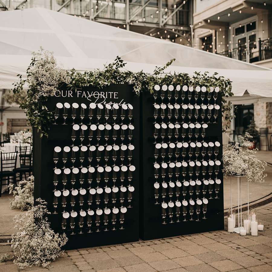 Black wedding champagne wall decorated with baby's breath, greenery, and candles in front of a tent