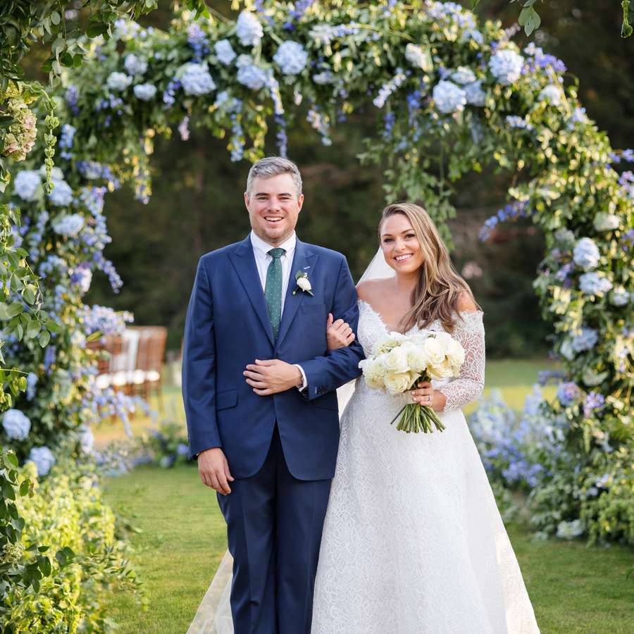 Bride and groom linking arms beneath a floral arch with blue hydrangea and delphinium and greenery 