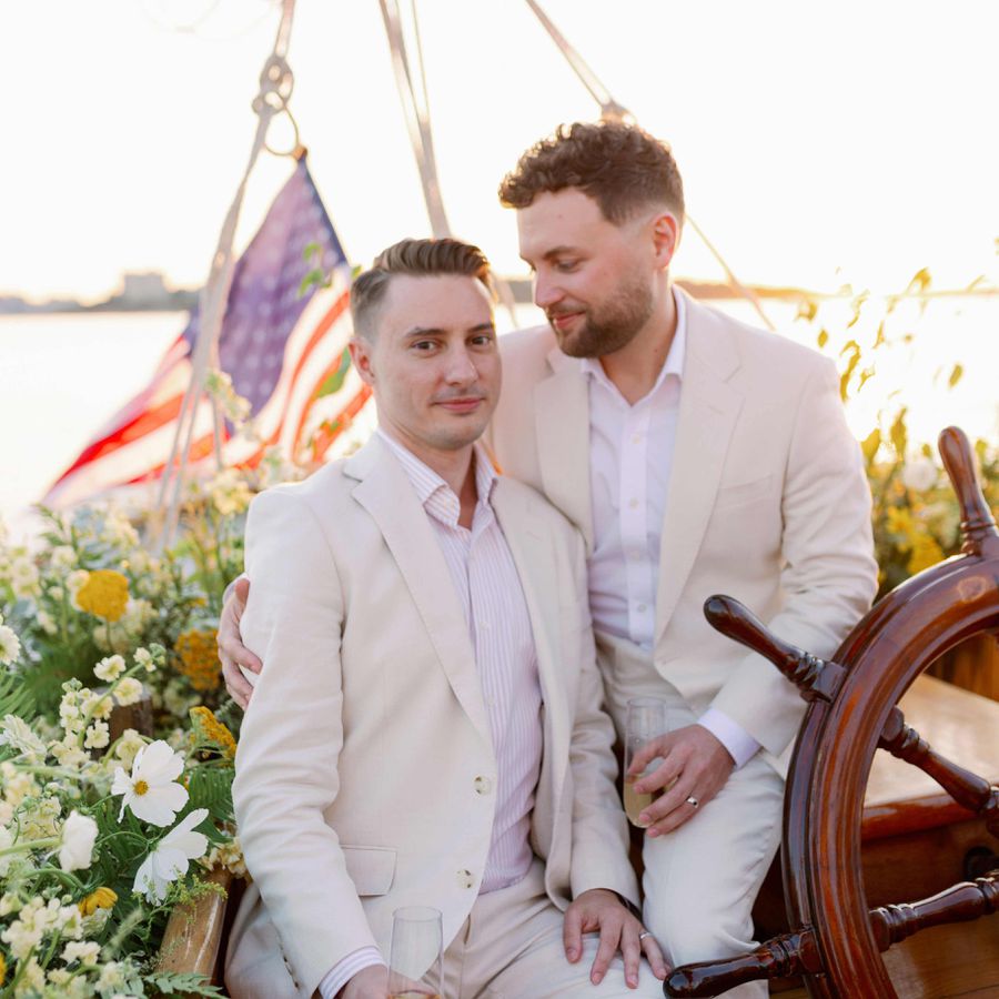 two grooms in cream-colored suits pose for a portrait on a schooner bedecked with white and yellow florals