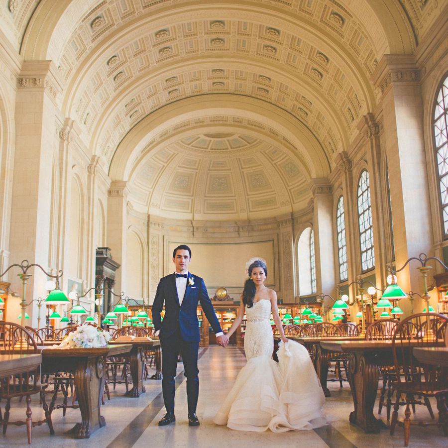 Bride and groom holding hands in the Boston Public Library, surrounded by wooden tables and green lamps during their wedding. 
