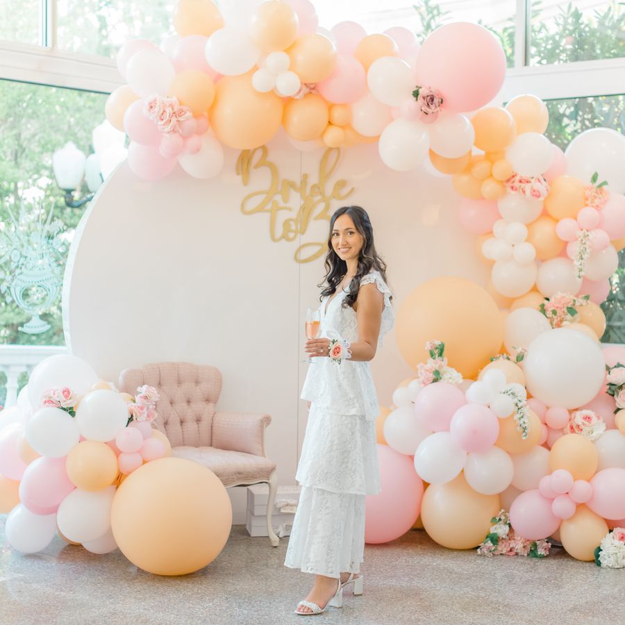 Bride in Tiered White Gown Standing in Front of Circular Bridal Shower Backdrop with Blush, White, and Orange Balloons