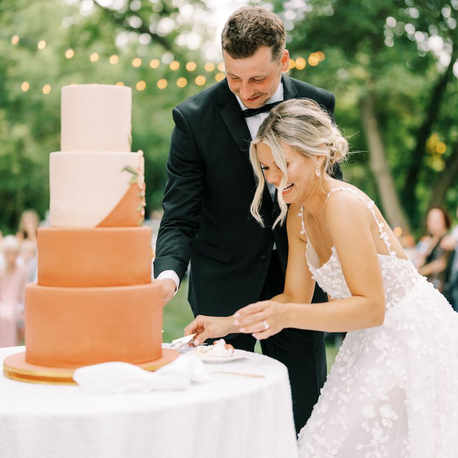 Bride and groom in formal wedding attire smiling and cutting a four-tier wedding cake at an outdoor wedding reception.