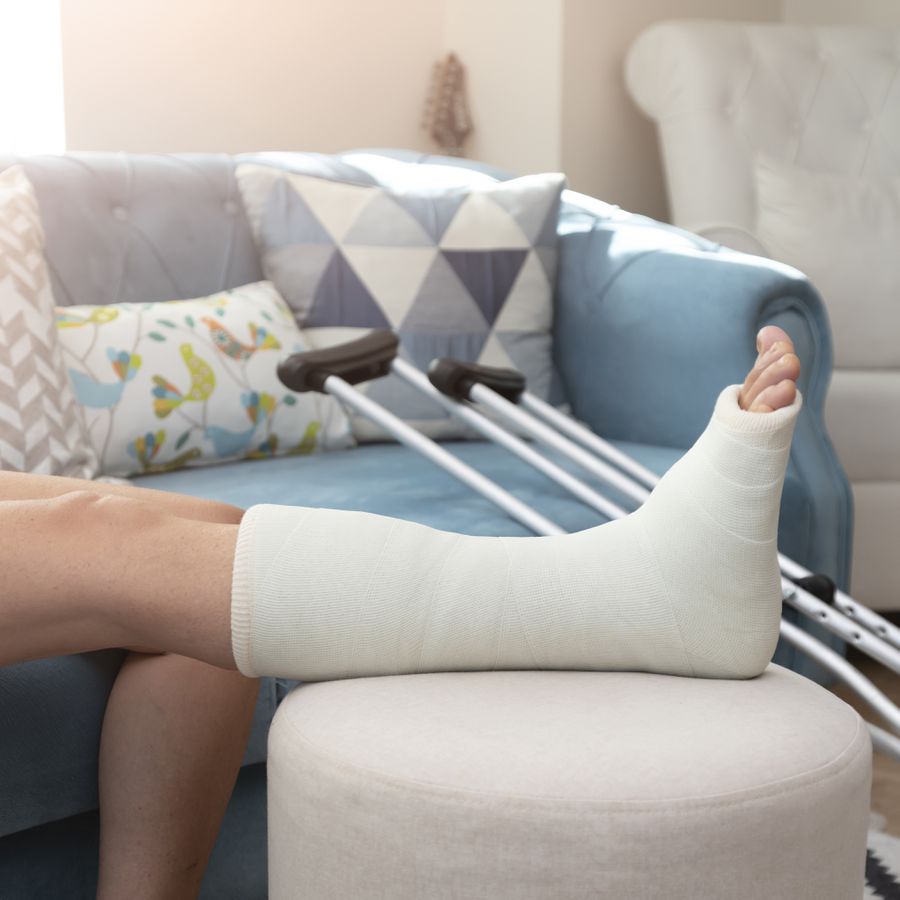 A personâs leg in a cast propped up on an ottoman with crutches lying on a blue couch in the background