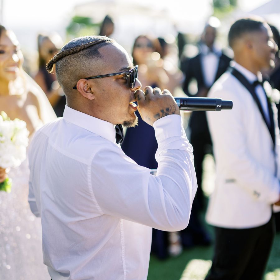 Bow Wow holding a microphone and signing at an outdoor reception with the bride and groom in the background