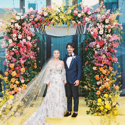 Bride in Floral Wedding Dress and Groom in Blue Suit Beneath Rainbow-Colored Floral Chuppah