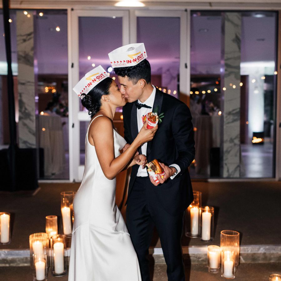 Bride in white gown and In-N-Out hat and groom in tuxedo and In-N-Out hat kiss while holding burgers