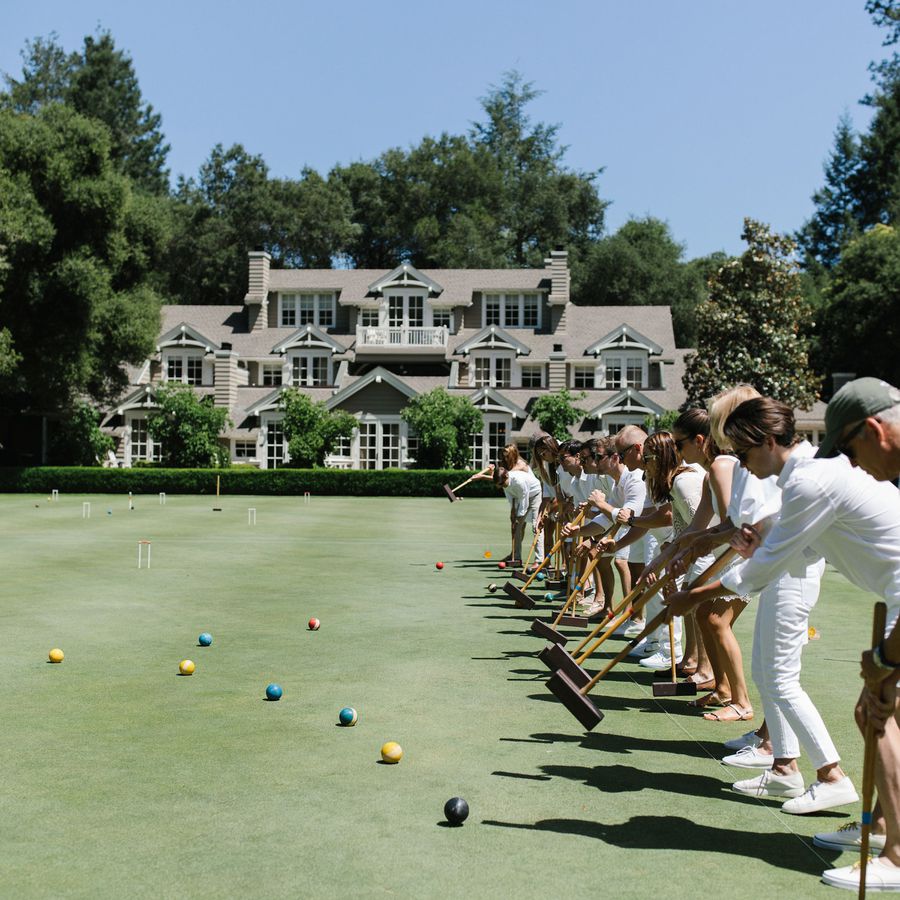 Members of a wedding celebration playing croquet on the lawn during a wedding reception in front of a large home on a sunny, summer day.