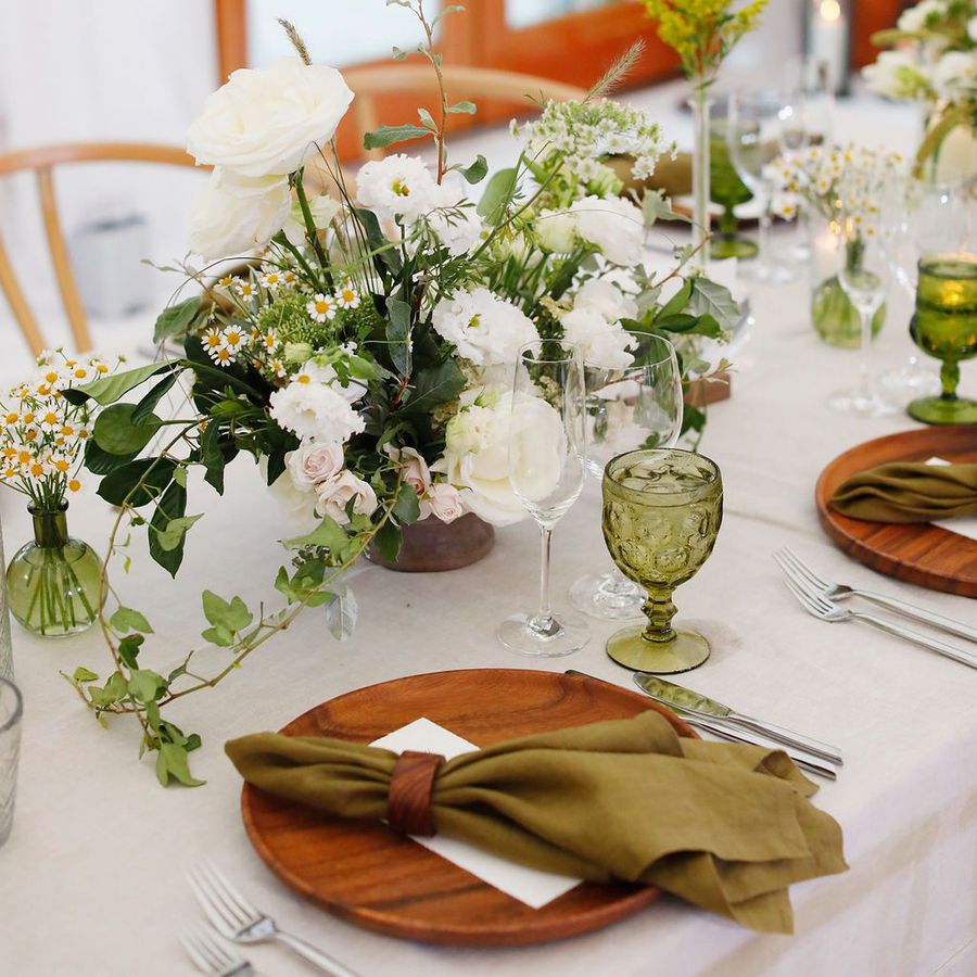 A tablescape with white linens, wooden chargers, green napkins, green goblets, and greenery centerpieces