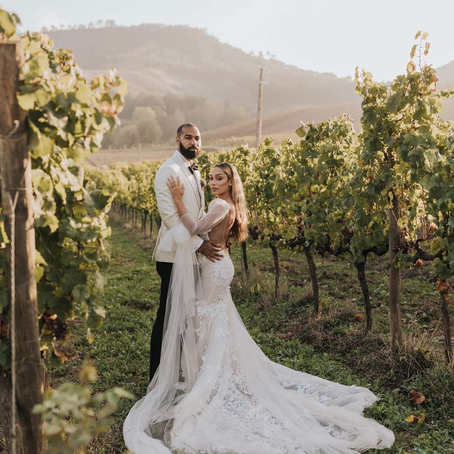 Groom in white suit jacket and bride in white long sleeve gown posing for portraits in a Portugal vineyard