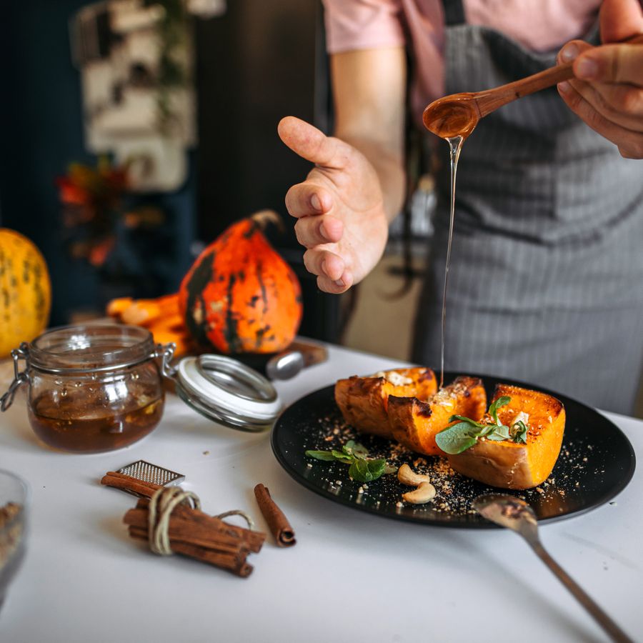 A chef pouring honey on top of roasted pumpkin dusted with cinnamon, which is placed on a table beside cinnamon sticks and other gourds