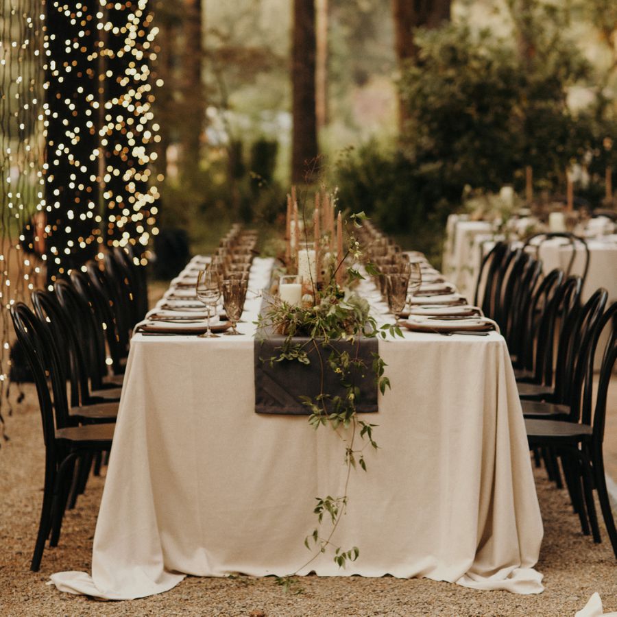 An outdoor forest wedding reception table with a white tablecloth, greenery, tapered candles, and hanging string lights.