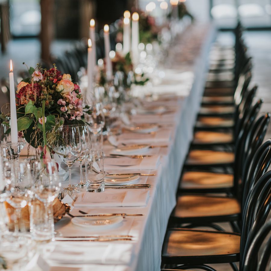 A wedding reception table decorated with tapered candles and fresh flowers, black chairs, glassware, and silver cutlery.