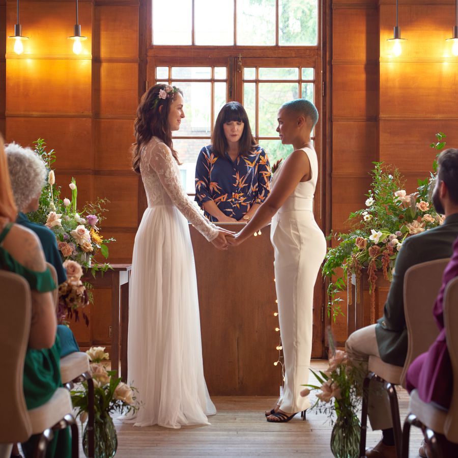 Two brides in white wedding attire exchange vows during a wedding ceremony officiated by a civil officiant. 