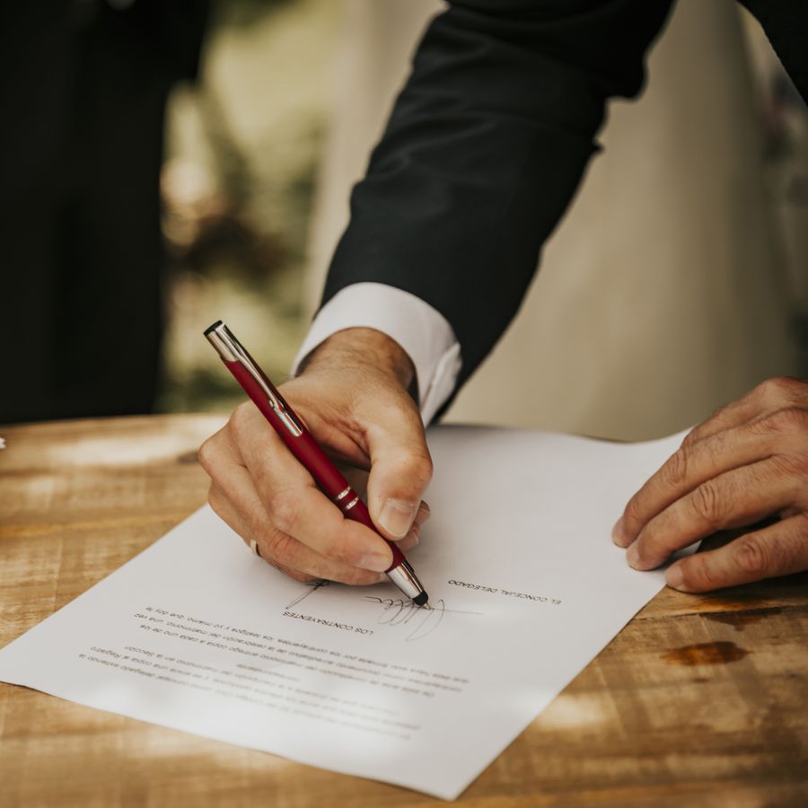 A groom in a black suit signing his marriage license with a red pen after the wedding ceremony.