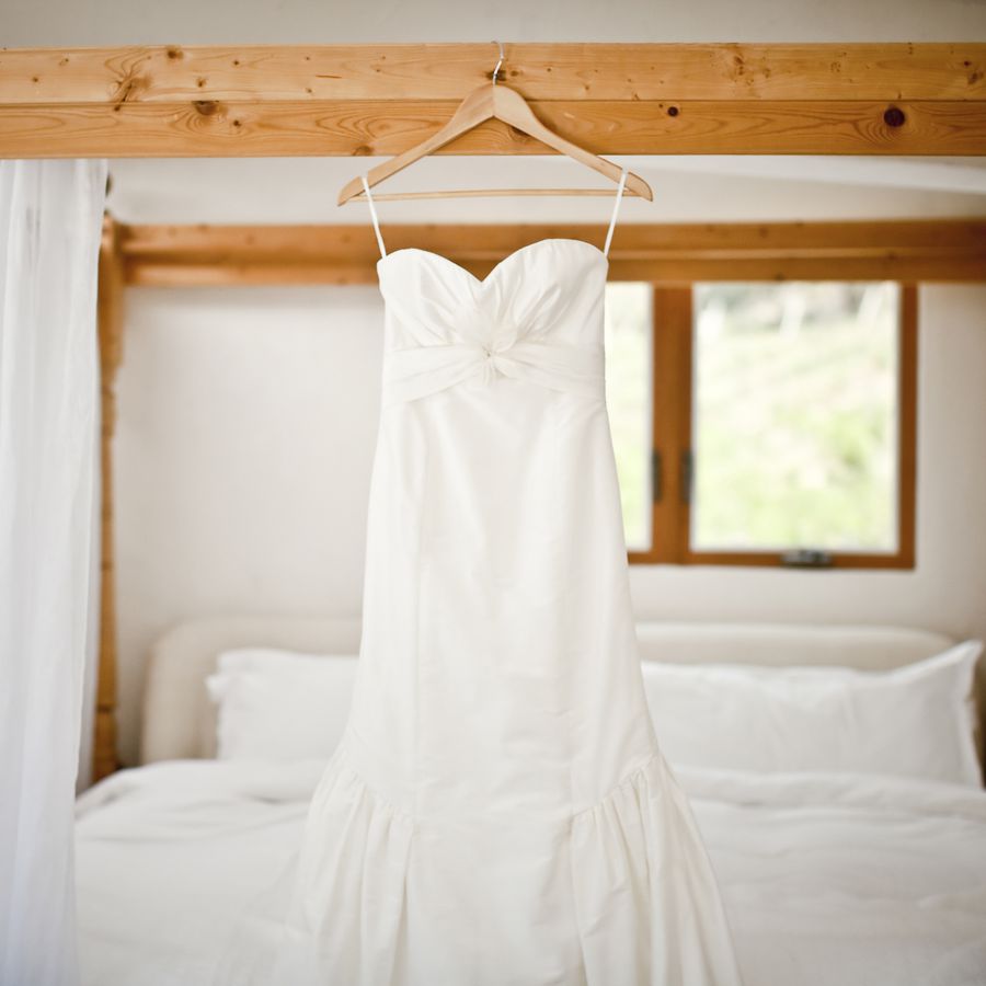 Strapless wedding dress suspended from a wooden hanger in a bedroom with white linens