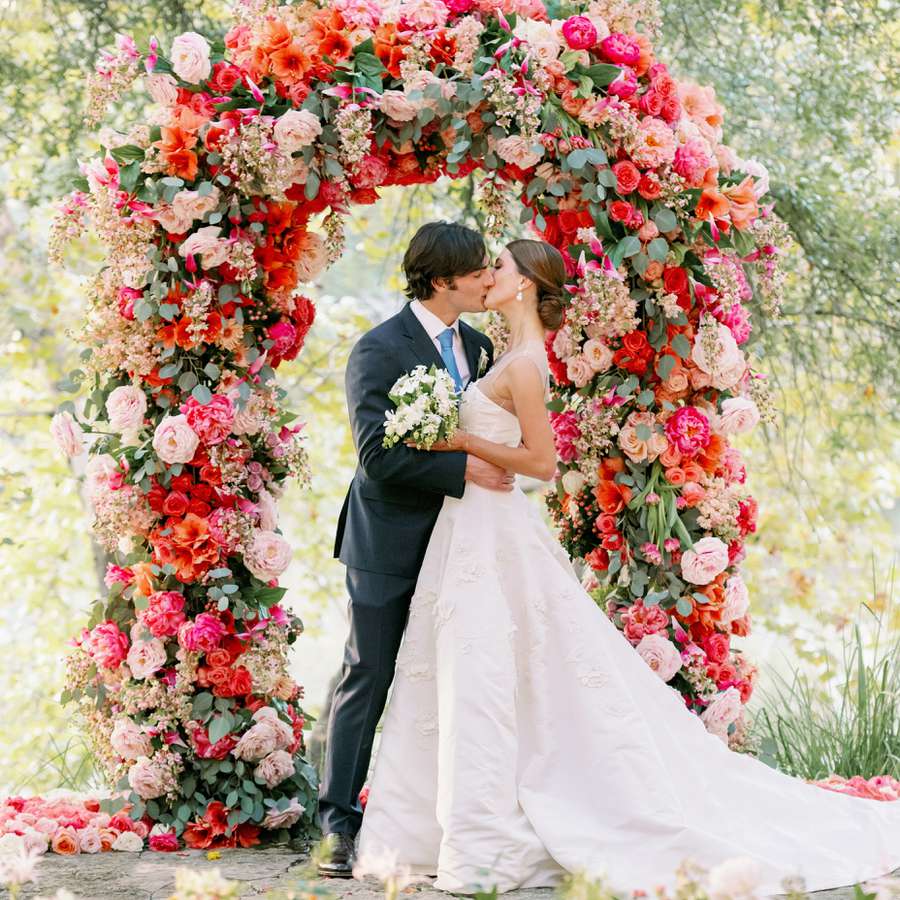 Bride and groom kiss in front of a floral arch with pink and red flowers and greenery