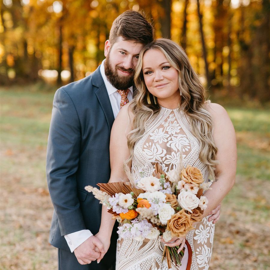 Groom with beard in suit and orange tie and bride with hair down and patterned halter dress holds a bouquet with orange, white, and purple florals