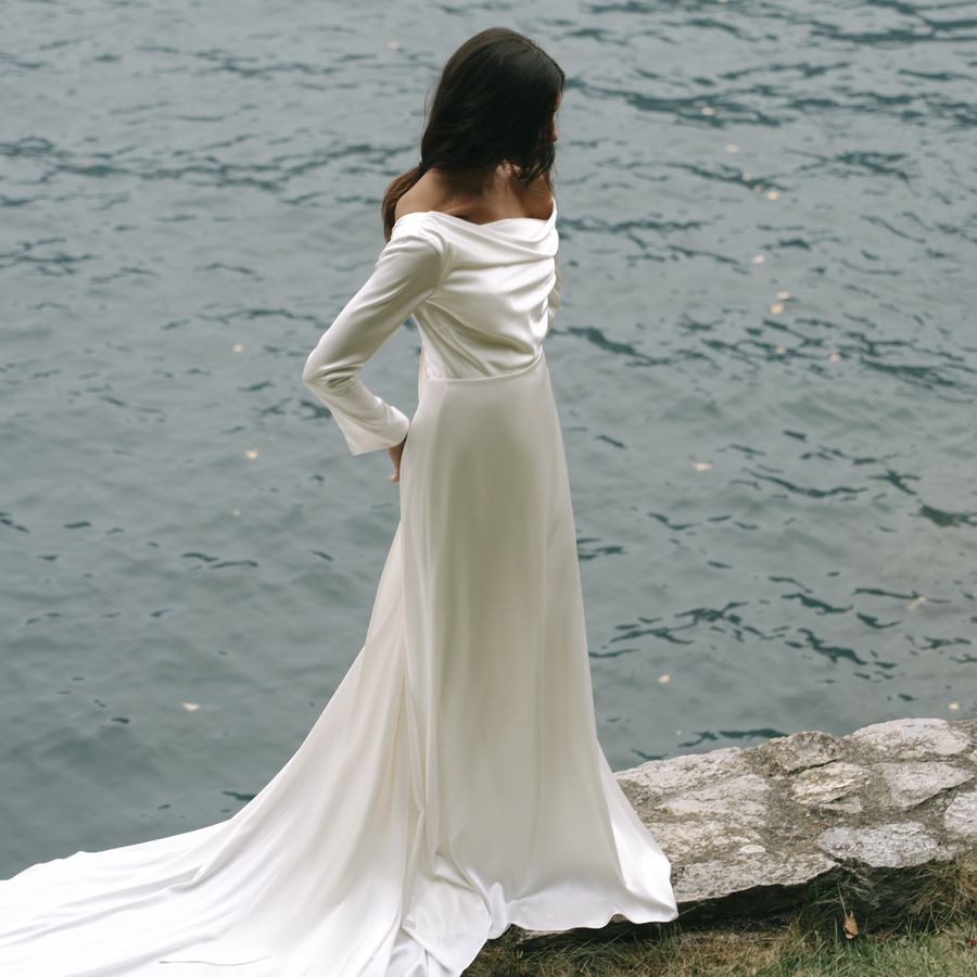 A bride wearing a silk off-the-shoulder long sleeve wedding gown, standing by the ocean.