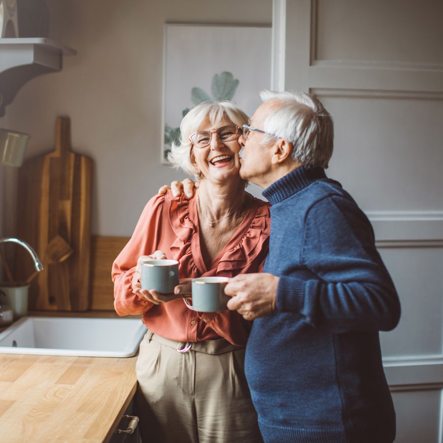 older couple drinking coffee in kitchen and kissing each other on the cheek