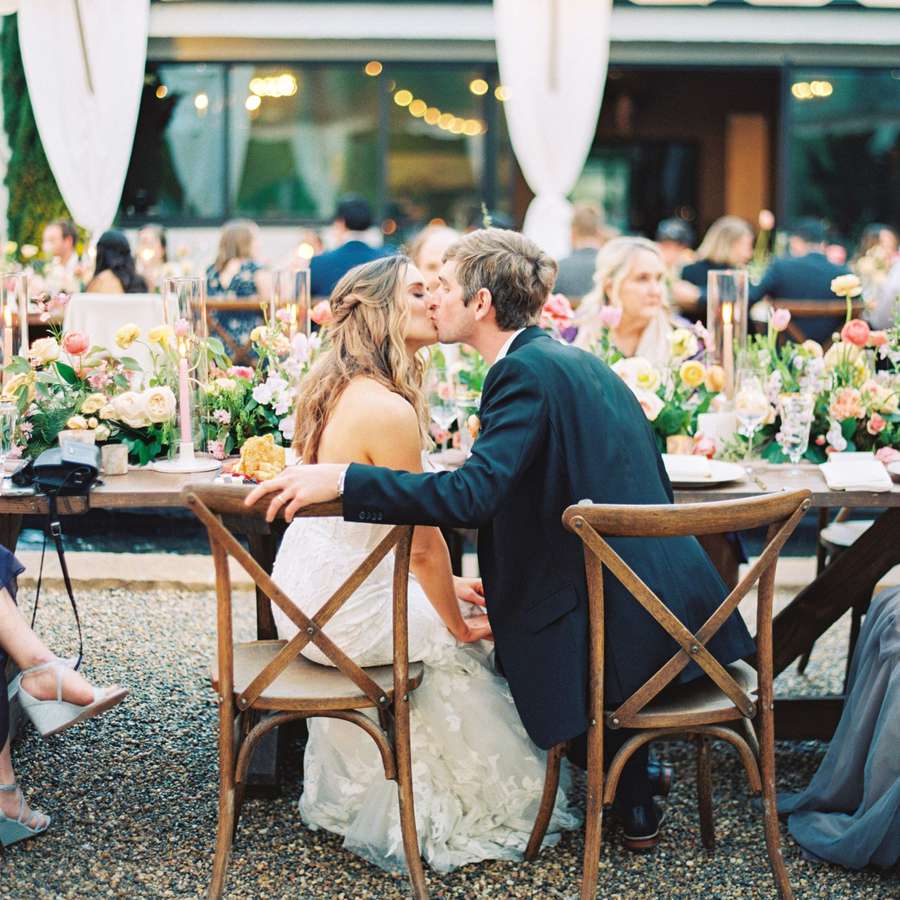 bride in whtie gown and groom in black suit kissing at wedding reception head table during a sumer wedding