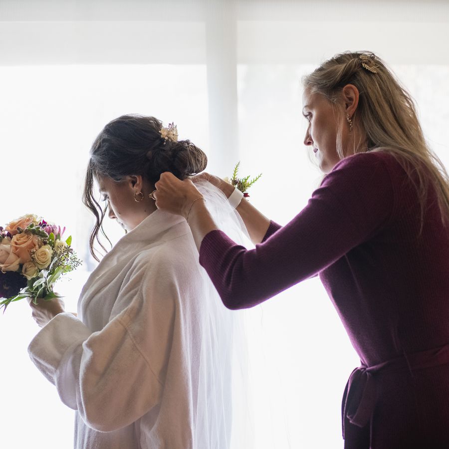 A personal attendant at a wedding helps the bride adjust her veil while getting ready. 