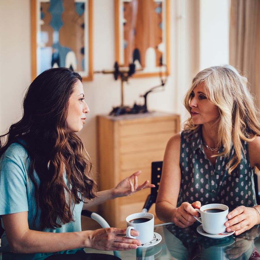 A mother and adult daughter talk while having coffee at home.
