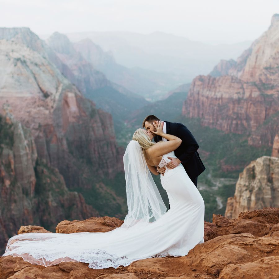 A bride in a white wedding gown and veil kisses her groom on top of a mountain at a national park.