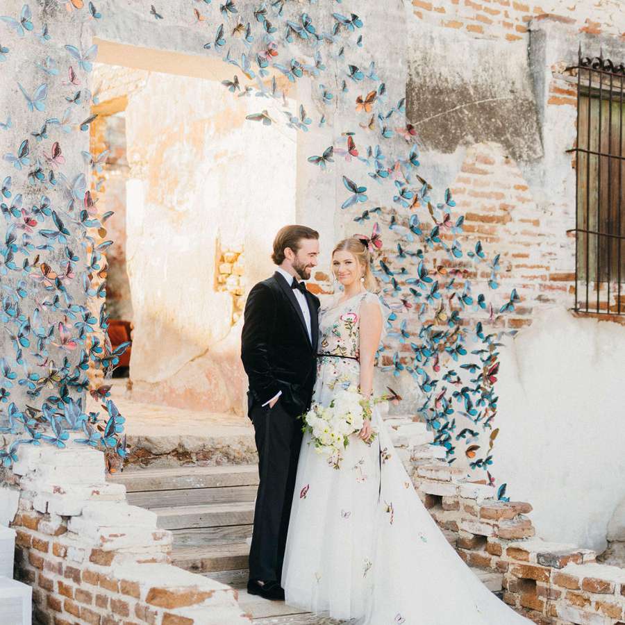 Bride in floral embroidered white gown and groom in tuxedo on steps with colorful butterfly installation