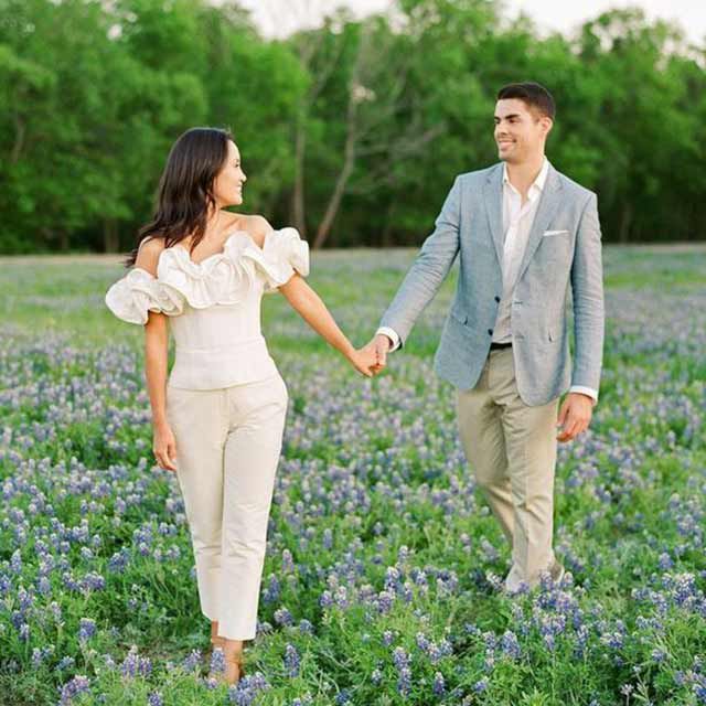 Bride in white ruffle top and pants and groom in gray suit walk in field of purple flowers