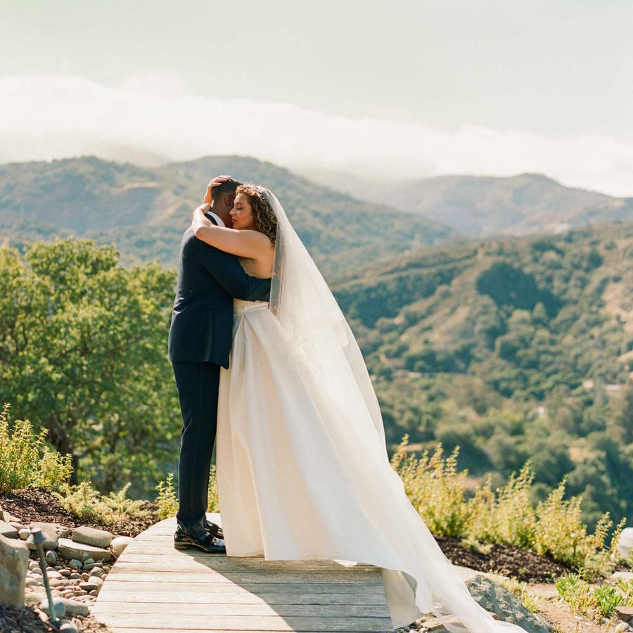bride and groom embrace on cliff with mountains in background