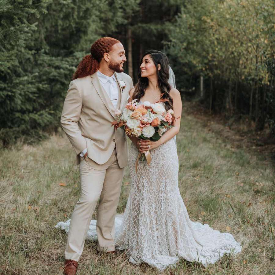 groom in tan suit and bride holding bouquet in field