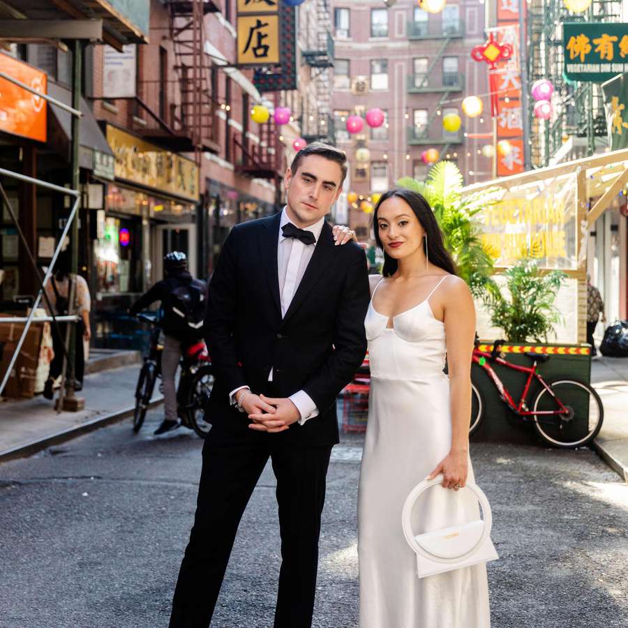 Groom in tuxedo and bride in white slip dress holding white purse on streets of New York City's Chinatown