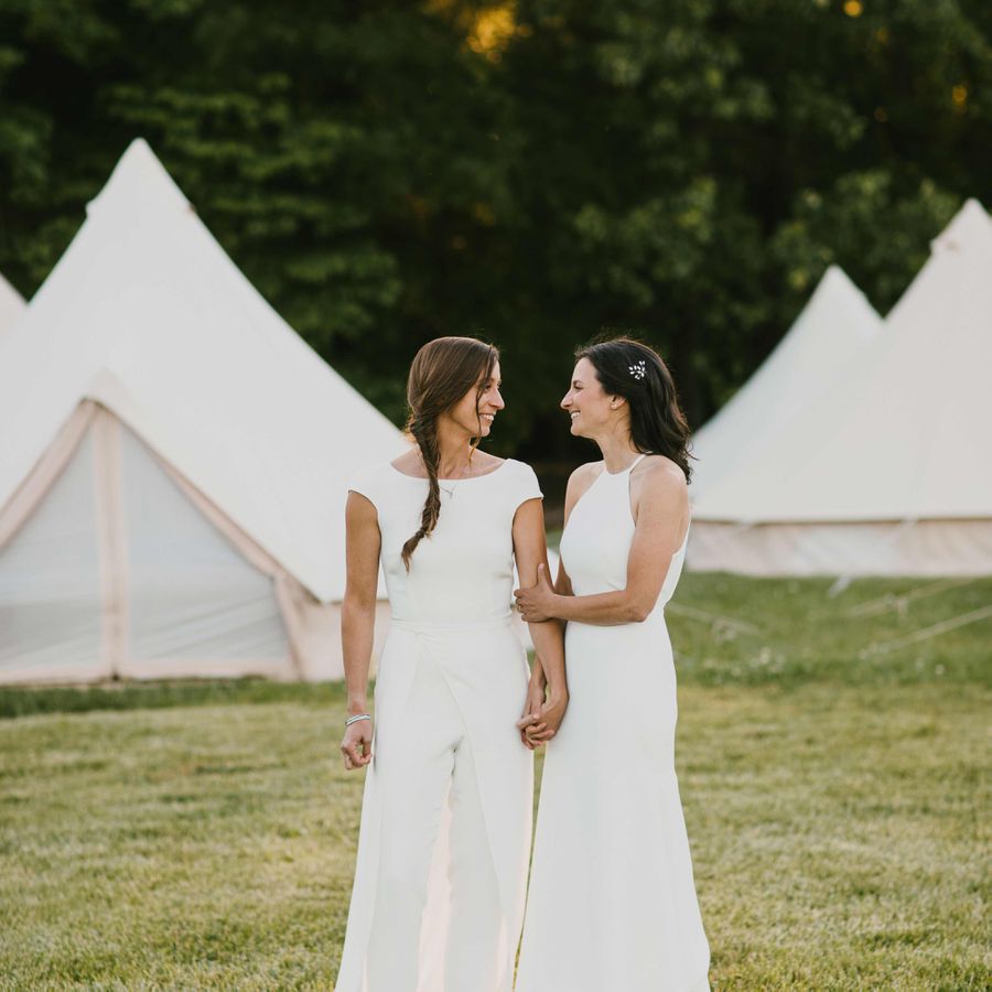 Bride with braid in white jumpsuit and bride with hair down in high-neck gown in front of camping tents