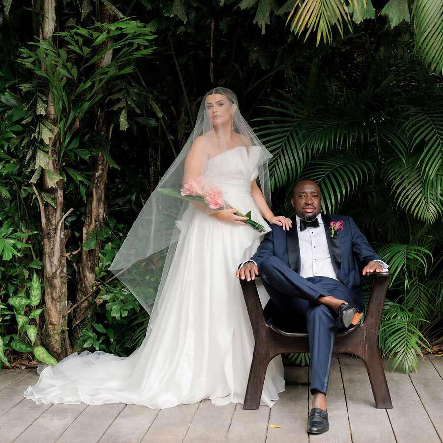 Bride in strapless gown and veil carrying pink bouquet next to groom in navy tuxedo sitting in chair in front of tropical plants
