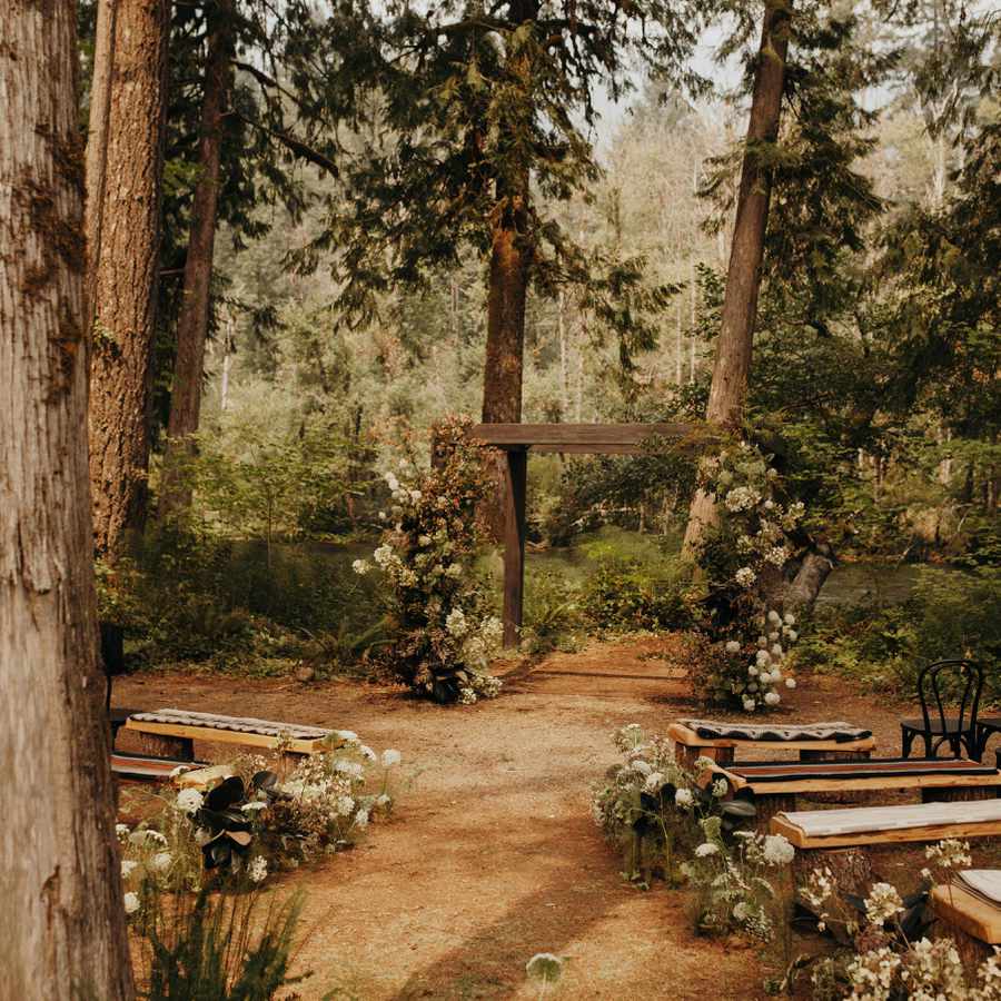 Forest wedding ceremony set up with wooden arch with greenery and white flowers, wood benches with blankets, and florals lining the aisles