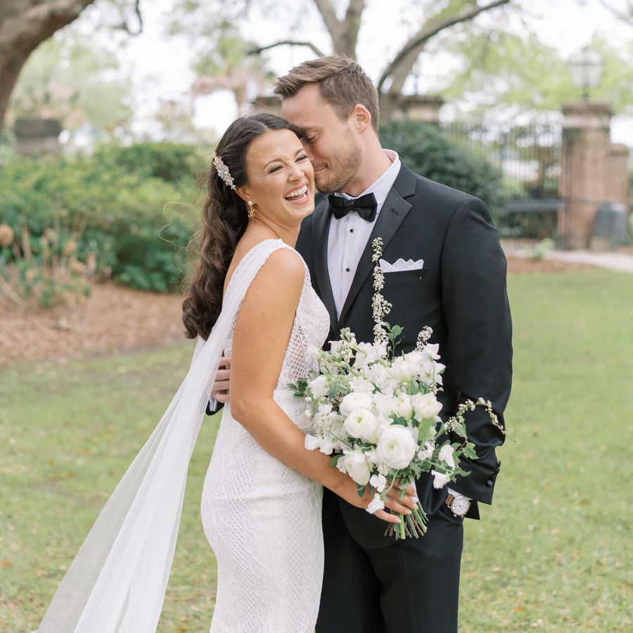 Bride in white sleeveless gown with train and hair pinned back holding a white bouquet with groom in black tuxedo outside
