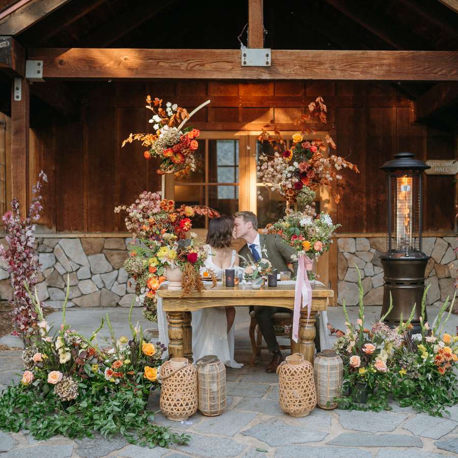 Couple kissing at a sweetheart table surrounded by flowers and rattan lanterns
