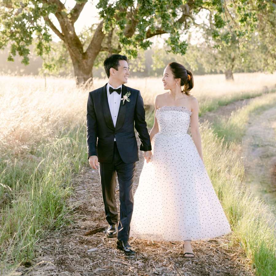 Groom in Black Tuxedo and Bride in Strapless Dotted Wedding Dress Walking in Meadow