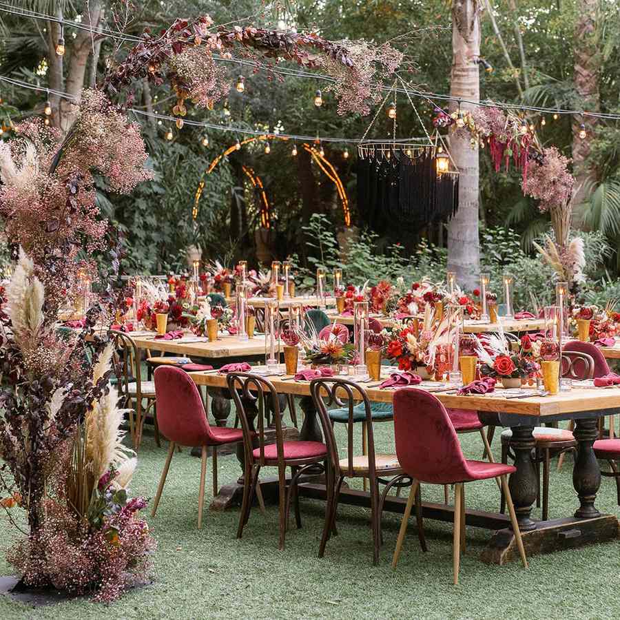 A reception setup with red velvet chairs tucked into wooden tables set with red napkins, red roses, and yellow glassware beneath a floral arch