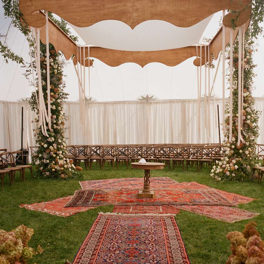 A tented ceremony setup with red patterned rugs, a wooden chuppah, and an aisle lined with antique hydrangeas
