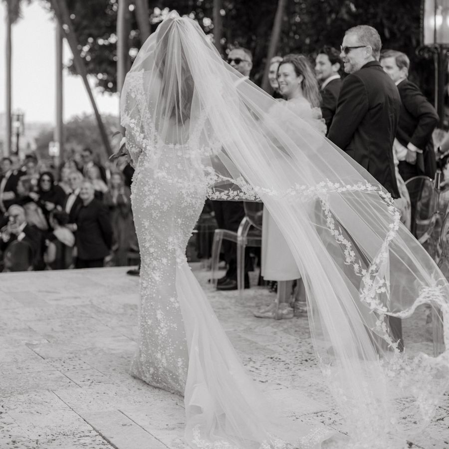 black-and-white image of bride walking down the aisle solo with an embroidered veil trailing behind her
