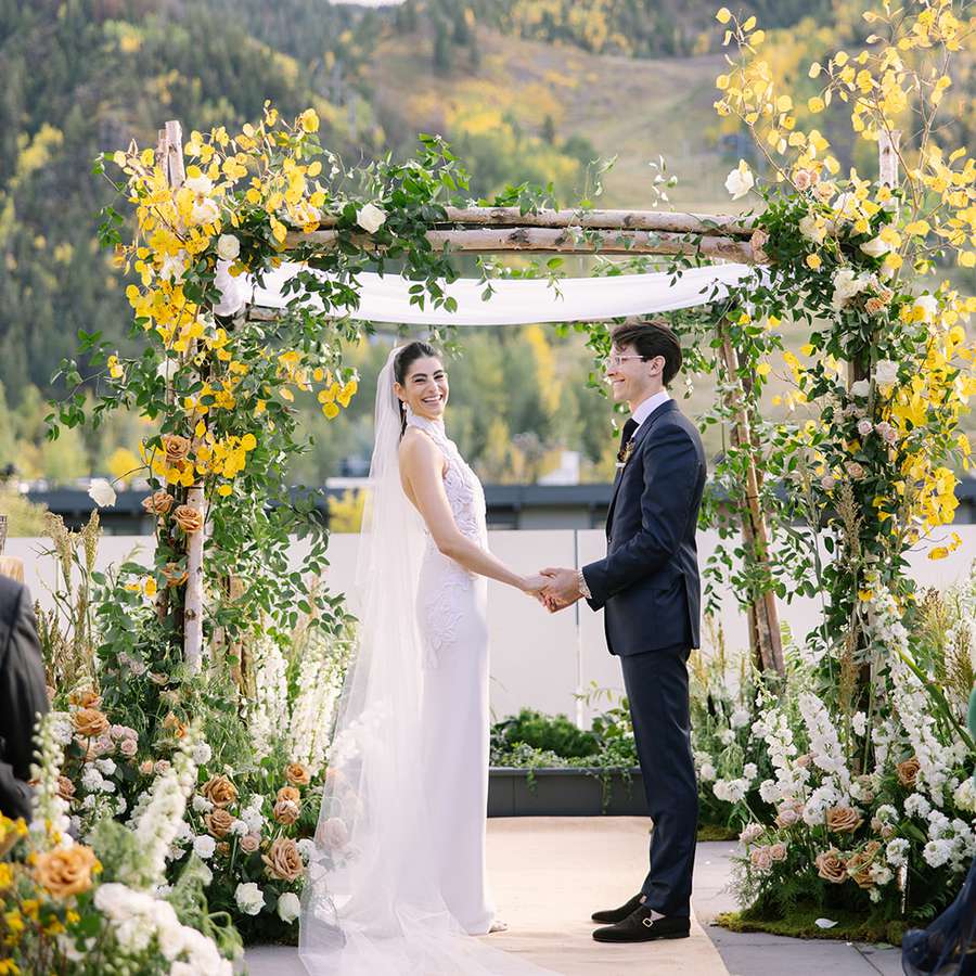 Bride and groom holding hands in front of a birch tree arch with gold leaves and white delphinium and toffee roses