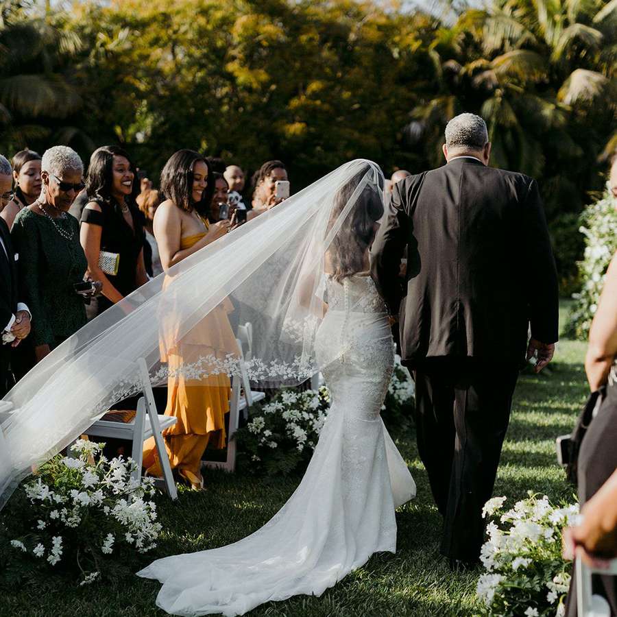 A bride wearing a white dress with a long veil walking with her father down the aisle at an outdoor wedding reception.