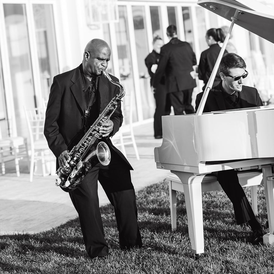 Black and white photo of saxophone player and piano player performing at an outdoor wedding reception.